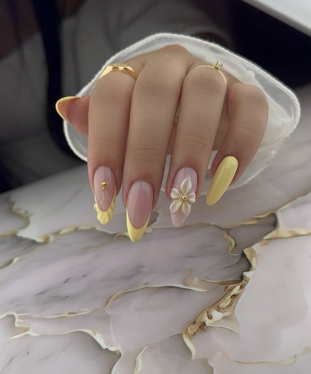 Long almond nails in pastel yellow and nude with a 3D white flower, gold beads and yellow French tips