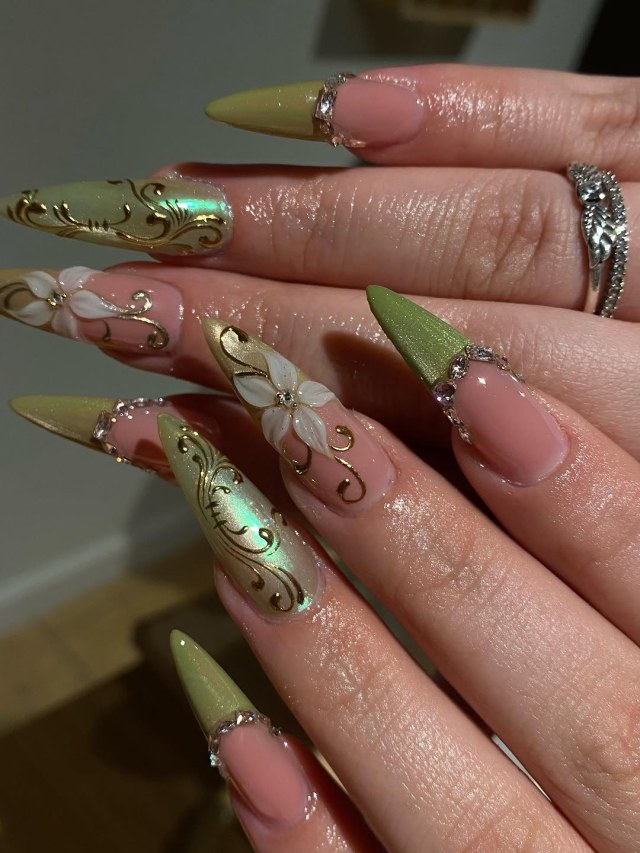 Extra-long stiletto nails with green chrome tips, gold swirl designs, 3D white flowers and rhinestone accents