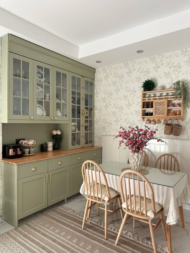 Sage green glass-front hutch with wood counter next to a dining table, rattan chairs and floral wallpaper